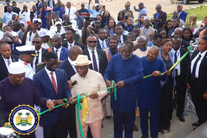 Group of dignitaries cutting a green ribbon at a formal ceremony as a large crowd looks on in the background.