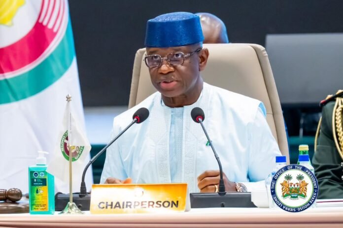 Chairperson at a formal meeting, speaking into two microphones with a CHAIRPERSON nameplate in front of him.