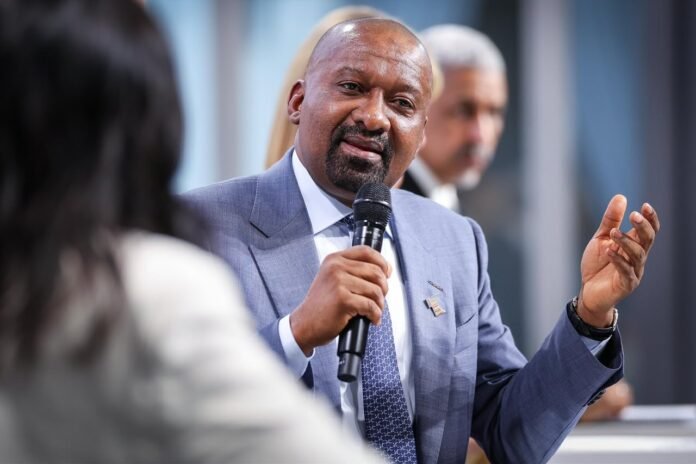Man in a blue suit speaks into a handheld microphone during a panel discussion, gesturing with his other hand, with a blurred figure in the background.