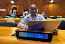 Sierra Leone Reaffirms Commitment to Climate-Resilient Urban Development and Inclusive Cities at UN General Assembly Man wearing glasses sits at a conference table with a laptop and a microphone, Sierra Leone nameplate in front, wood-paneled room behind him.