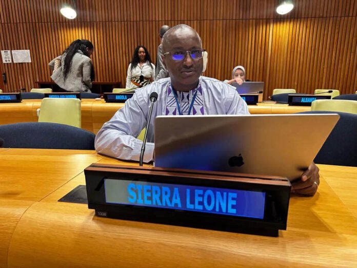 Man wearing glasses sits at a conference table with a laptop and a microphone, Sierra Leone nameplate in front, wood-paneled room behind him.