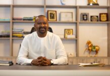 OP-ED: Fragility and the Politics of Prevention Man in a white traditional outfit sits at a desk in an office, smiling, with awards and books on shelves behind him.