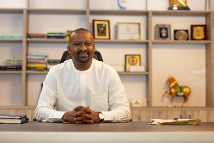 Man in a white traditional outfit sits at a desk in an office, smiling, with awards and books on shelves behind him.