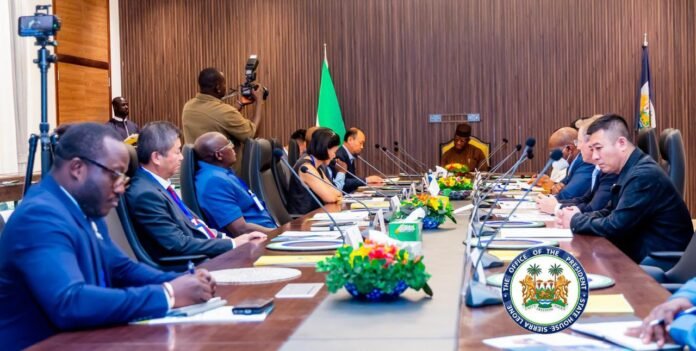 Dignitaries seated along a long conference table in a formal meeting, with flags behind them and a presidential seal in the foreground.