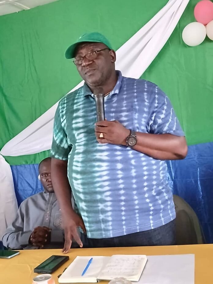 Man in a green cap and blue tie-dye shirt speaks into a handheld microphone at a table with notebooks and phones, colorful backdrop behind him.