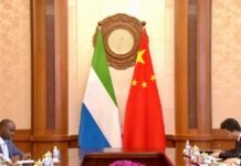 Fifty-Five Years Side by Side: The Resilience and Warmth of China–Sierra Leone Friendship In a formal meeting room, two flags stand behind a carved clock: a green, white, and blue tricolor on the left and a red flag with yellow stars on the right, with people seated at a conference table in the foreground.