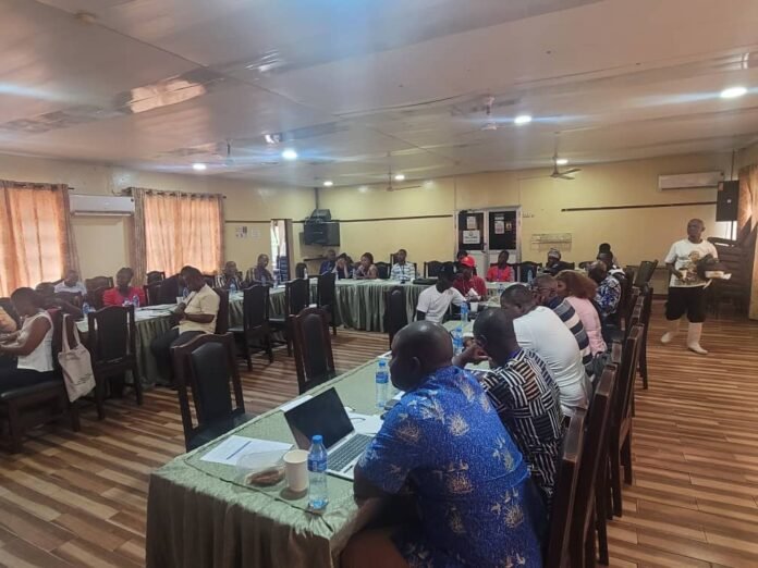 People seated around U-shaped conference tables in a meeting room, some with laptops and notebooks, a speaker standing at the back right.