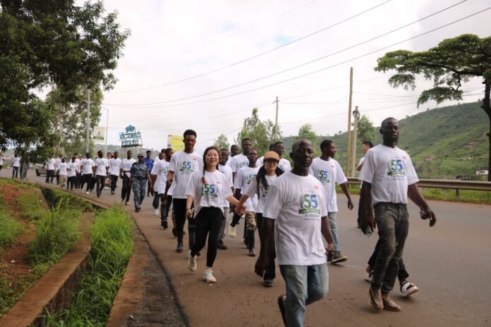 Group of people walking together on a paved road for a celebration, all wearing white T-shirts with a 55th anniversary design, countryside backdrop with hills and trees