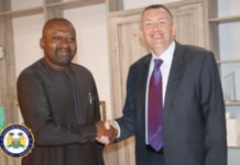 Vice President, Trócaire CEO Discuss Longstanding Partnership With Sierra Leone Two men shake hands indoors: one in a dark traditional outfit, the other in a suit, with a government-style seal in the bottom-left corner.
