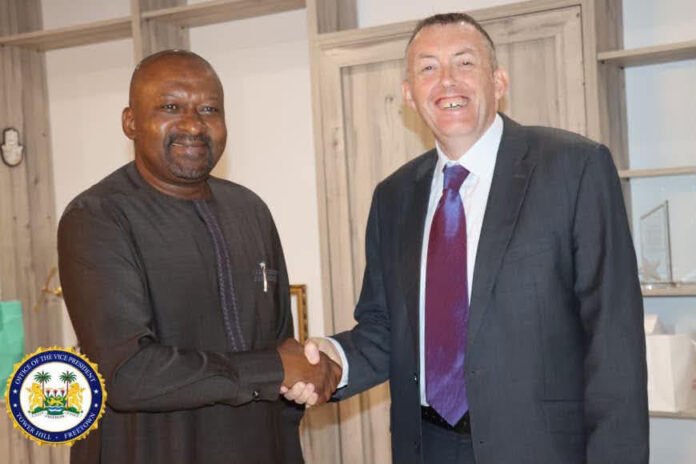 Two men shake hands indoors: one in a dark traditional outfit, the other in a suit, with a government-style seal in the bottom-left corner.
