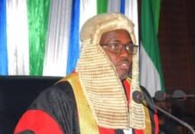 Government and Opposition Locked in Debate as Constitution Amendment Bill Advances Judge in a white barrister wig and red-black robes speaks at a microphone during a formal ceremony with colorful backdrop.
