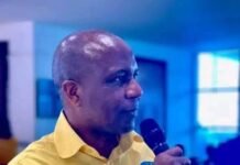 NASSIT Deputy Director General Hails Outgoing Director General’s Transformational Legacy Man in a yellow shirt speaks into a handheld microphone at an indoor event, with blurred attendees in the background.