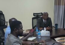 Lands Minister Calls for Stronger Controls on Illegal Building and Urban Development Three men sit around a large conference table in a meeting room, one in a suit speaking while others listen, with water bottles and tissues on the table.