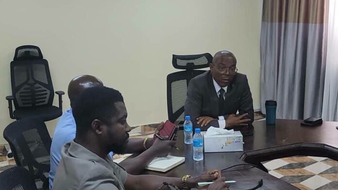 Three men sit around a large conference table in a meeting room, one in a suit speaking while others listen, with water bottles and tissues on the table.