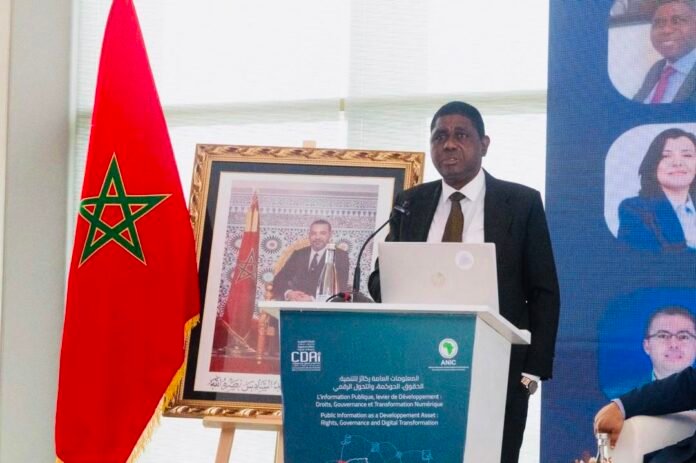 Speaker in a dark suit delivers a talk at a podium with a laptop, Moroccan flag to the left, and a framed portrait behind.