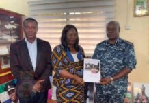 IGP Sellu Commits to Implementing UNDP Recommendations on Family Support Unit Three adults pose for a formal presentation: a man in a suit, a woman in a patterned dress, and a uniformed officer holding a printed document in a room with posters in the background.
