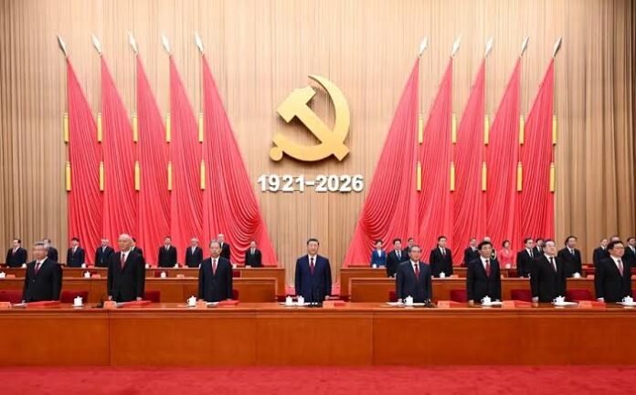 Official ceremony with a large gold hammer-and-sickle emblem and the dates 1921–2026, surrounded by red banners and officials at a long dais.