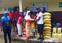 Jorg Wolf Foundation Distributes €10,000 Worth of Relief to Vulnerable Communities Across Sierra Leone People at a charity distribution outside a building: a man hands a plate to a girl while by stacked rice sacks, oil containers, and boxes; volunteers and recipients watch.
