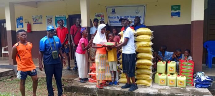 People at a charity distribution outside a building: a man hands a plate to a girl while by stacked rice sacks, oil containers, and boxes; volunteers and recipients watch.