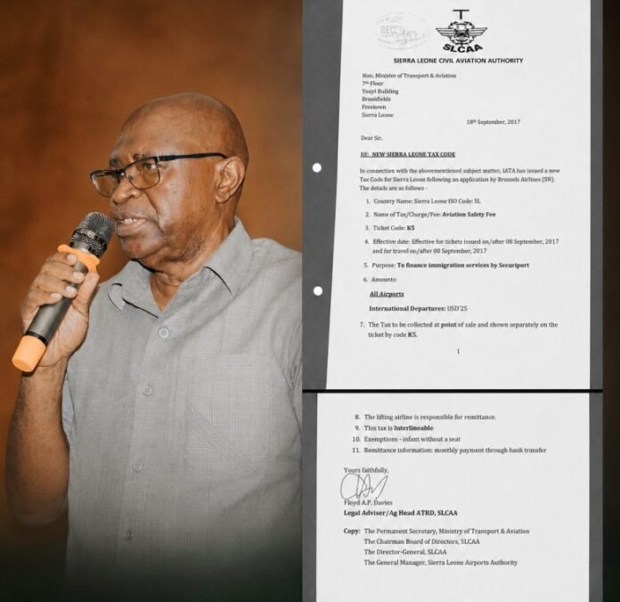 Older man wearing glasses and a grey shirt speaks into a handheld microphone beside a large printed document in a binder.