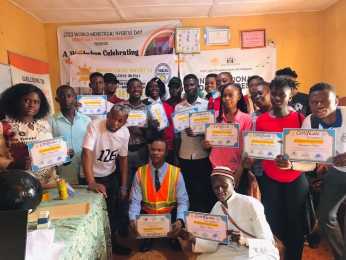 Group of people posing with certificates at a community awards event for World Menstrual Hygiene Day, banners visible in the background.