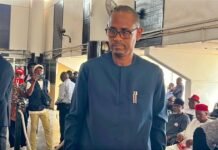 Chericoco Emerges as Leading Contender for APC’s 2028 Presidential Flagbearer Race Man in a blue traditional outfit with glasses walking through a crowded indoor event, with attendees seated in the background.