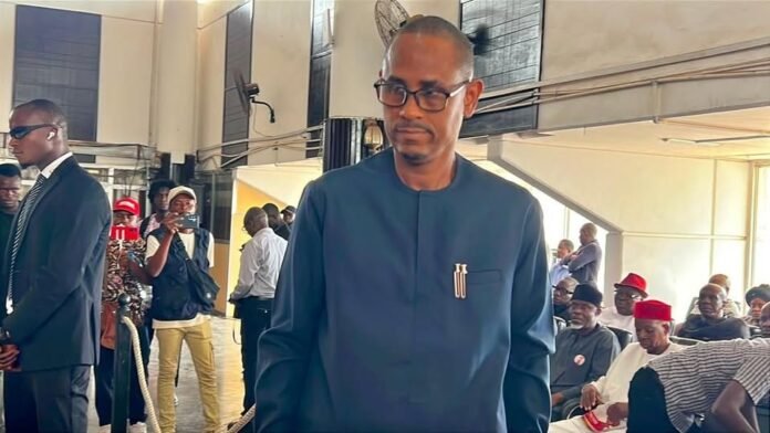 Man in a blue traditional outfit with glasses walking through a crowded indoor event, with attendees seated in the background.