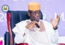 Sierra Leone Gears Up for Major ECOWAS Summit Under President Bio’s Leadership Man in traditional white robe and tan cap sits in a red chair, speaking into a handheld microphone at a formal event.