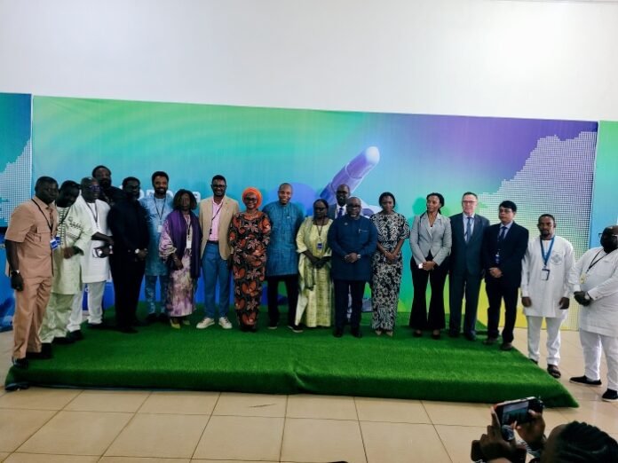 Group of men and women in formal and traditional attire posing on a green stage in front of a colorful backdrop at a conference event.