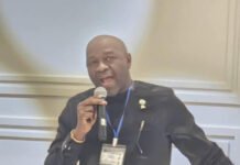 Dr. Gilpin Highlights Digital Trust and Financial Inclusion at SLCA 2026 in Canada Man in a black traditional outfit speaks into a handheld microphone at a conference, wearing a blue badge lanyard.