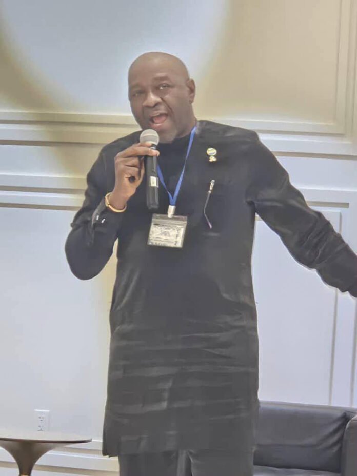Man in a black traditional outfit speaks into a handheld microphone at a conference, wearing a blue badge lanyard.