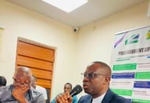 NPPA Unveils New Salone e-GP Modules to Boost Transparency, Efficiency and Service Delivery Man in a navy suit speaks into a handheld microphone at a conference table, with a notepad and phone in view, during a meeting.