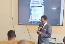 YouBest, Connaught Hospital and Gemstar Pharmacy Disclose Breakthrough Diagnostic Solutions A man in a gray suit giving a presentation in a classroom, speaking into a microphone while reading from notes as a large screen shows a conference poster titled Internal Medicine–Chemical Pathology.