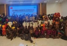 China and Sierra Leone Celebrate 55 Years of Diplomatic Relations, Renew Commitment to Stronger Bilateral Cooperation Large, diverse group of people in colorful traditional clothing posing for a group photo at a cultural event, with a banner on a screen behind them.