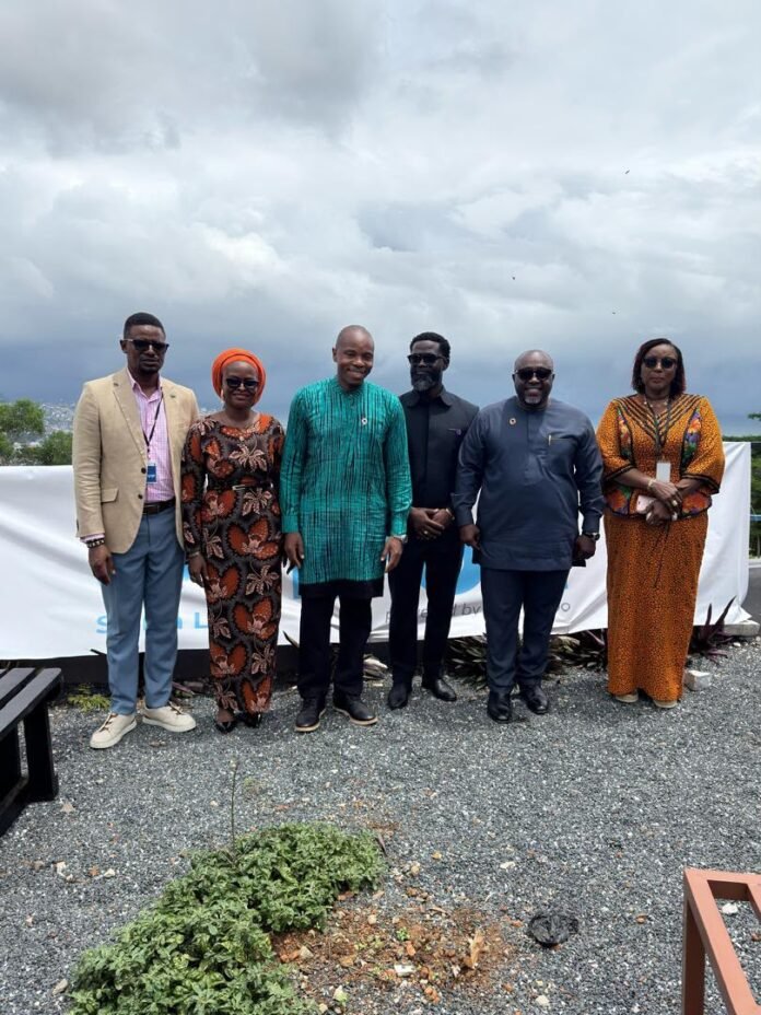 Six people stand in a row on a rooftop, wearing colorful traditional African outfits under a cloudy sky – group portrait.