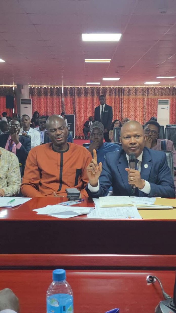 Man in a blue suit speaks into a microphone beside a man in orange traditional attire at a conference panel, with audience seated behind them.