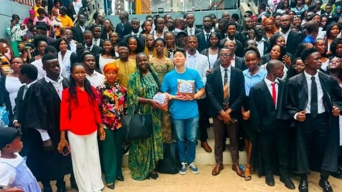 Large diverse group of graduates and attendees posing for a photo on a staircase, with a person in a blue shirt holding books at the center.