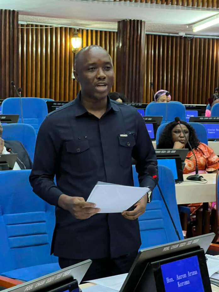 Man in a dark shirt speaks in a formal meeting room, holding a white folder with blue seats and microphones in the background.