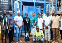 Mayor Aki-Sawyerr Inspects New Water Kiosks and Public Toilets to Boost WASH Services Across Freetown Group of community volunteers posing in front of a blue water-tank facility and chain-link fence.