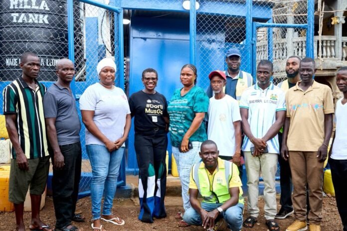 Group of community volunteers posing in front of a blue water-tank facility and chain-link fence.