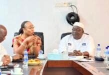 NCRA Assures APC, Moral Guarantors of Credible Civil and Voter Registration Process Three people sit around a conference table, a woman in patterned dress speaking while others listen. Water bottles and snacks are on the table.