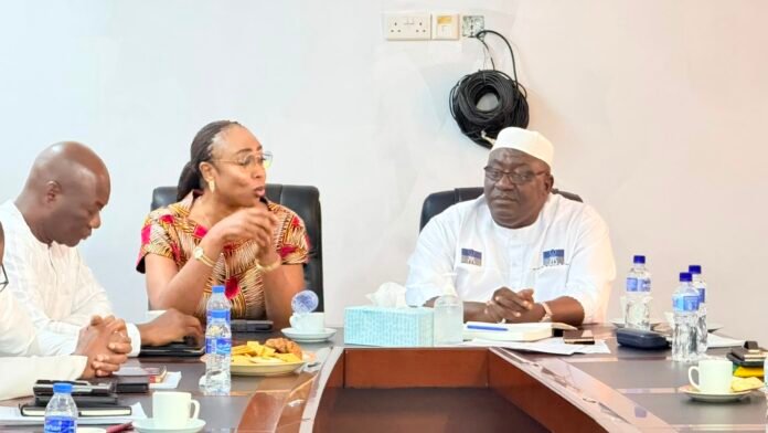 Three people sit around a conference table, a woman in patterned dress speaking while others listen. Water bottles and snacks are on the table.
