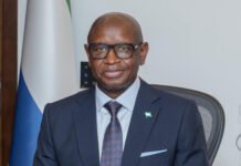 President Bio Signals Possible Clemency for Former President Koroma as ECOWAS Summit Opens Smiling man in a navy suit and glasses sits at a desk with a flag and official seal in the background, appears to be in a formal office.