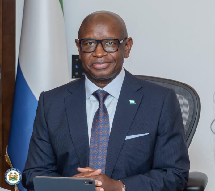 Smiling man in a navy suit and glasses sits at a desk with a flag and official seal in the background, appears to be in a formal office.
