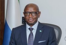 President Bio Grants Clemency to Former President Koroma as ECOWAS Summit Gets Underway Professional man in a dark navy suit and glasses sits at a desk in an office, with a flag in the background.