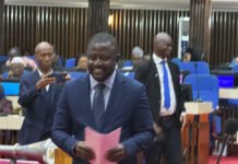 Attorney General Lays African Human Rights Court Protocol Before Parliament Smiling man in a navy suit holding pink notes at a formal meeting or ceremony in a conference room; attendees in the background.