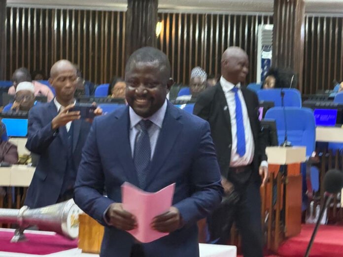 Smiling man in a navy suit holding pink notes at a formal meeting or ceremony in a conference room; attendees in the background.