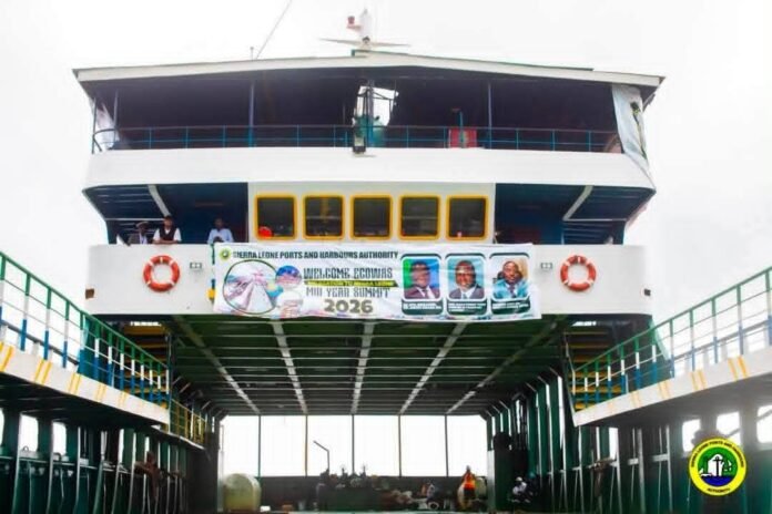 Two-level white ferry docked at a pier, with yellow-framed windows and a banner across the front deck and lifebuoys on both sides.
