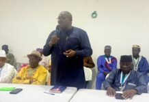 NCRA DG Commends Tribal Heads for Strengthening Civil Registration Across Sierra Leone Man in dark blue attire stands and speaks into a microphone at a meeting, surrounded by seated attendees in traditional outfits.