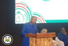 VP Hails President Bio’s Legacy at Historic Commissioning of Julius Maada Bio Conference Centre & ECOWAS Logistics Depot Man in a blue traditional outfit speaks at a wooden podium on a stage, with a large screen showing colorful circular graphics behind him and an official seal visible on the left.